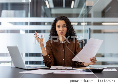 Businesswoman feeling stressed and overwhelmed, sitting at her office desk with laptop and documents, experiencing confusion and frustration with a complex work challenge 133182074