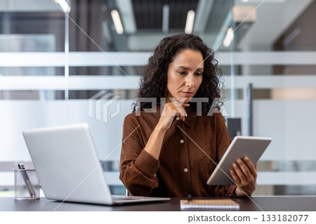 Professional businesswoman in smart casual attire reviewing data on a digital tablet at her desk with a laptop, focused and productive in a modern hybrid office workspace Professional businesswoman in smart casual attire reviewing data on a digital tablet at her desk with a laptop, focused and productive in a modern hybrid office workspace 133182077