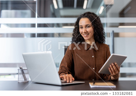 Young businesswoman with curly hair smiling and actively engaged in work, balancing a laptop and a tablet at her modern office desk, showcasing efficiency and productivity 133182081