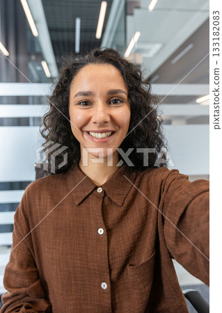 Smiling young woman with beautiful curly hair taking a selfie or participating in a video call, portraying concepts of remote work, communication, and happiness in an office environment 133182083