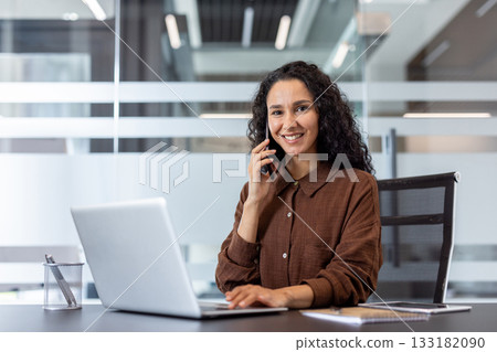 Young businesswoman communicating on a phone call, smiling while typing on her laptop at a modern office desk, demonstrating professionalism and productivity Young businesswoman communicating on a phone call, smiling while typing on her laptop at a modern office desk, demonstrating professionalism and productivity 133182090