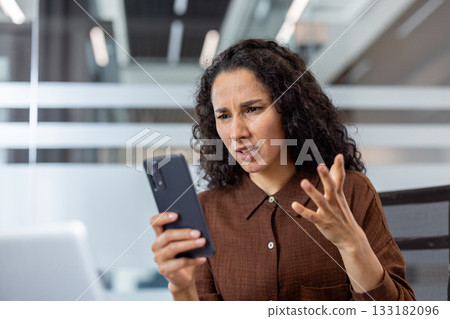 Young businesswoman with curly hair expressing anger and frustration while reading a message on her mobile phone, experiencing problems or bad news at her modern workplace 133182096