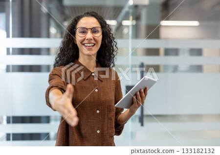 Professional businesswoman in a modern office smiling and reaching out for a handshake while holding a tablet, conveying welcome, partnership, hiring and confident collaboration Professional businesswoman in a modern office smiling and reaching out for a handshake while holding a tablet, conveying welcome, partnership, hiring and confident collaboration 133182132