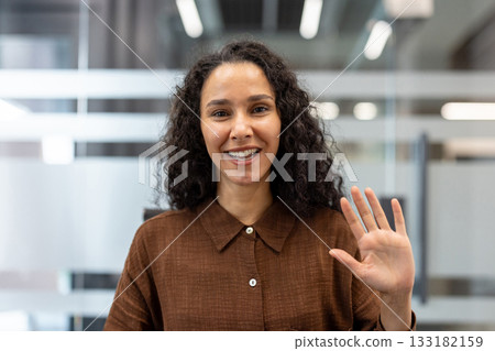 Woman with curly hair smiling and waving at camera during a remote video conference from a modern office workspace, friendly professional connecting virtually with colleagues Woman with curly hair smiling and waving at camera during a remote video conference from a modern office workspace, friendly professional connecting virtually with colleagues 133182159