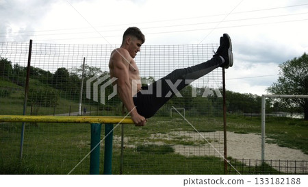 Strong man trains abdominal muscles raising legs on parallel bars at sports ground. Young shirtless sportsman exercising at nature. Sporty athlete working out at nature. Concept of sportive lifestyle 133182188