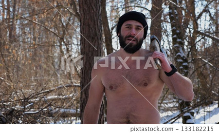Portrait of shirtless bearded man stands in a snowy winter forest with axe. Tired guy looking into camera after chopping logs in snowy woodland. The scene represents outdoor work in cold condition 133182215