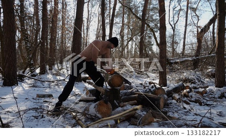 Shirtless man sawing a log in sunny winter forest. Strong guy working with saw in snowy woodland. The scene highlights human effort and outdoor activity in cold conditions. Slow motion 133182217