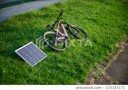 Pink electric mountain bike lies on grassy lawn, connected to solar panel for charging. Bike's rugged design amidst vibrant green grass, with paved pathway nearby. Pink electric mountain bike lies on grassy lawn, connected to solar panel for charging. Bike's rugged design amidst vibrant green grass, with paved pathway nearby. 133183171