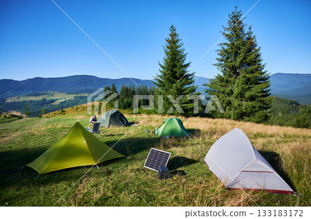 Tourist tents with solar panel and portable power station nearby in grassy field. Scene surrounded by rolling hills, tall pine trees under clear blue sky, showcasing eco-friendly camping setup. 133183172
