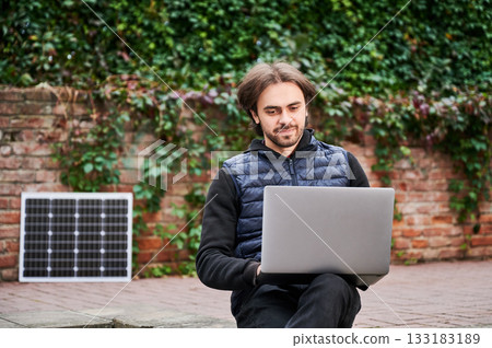 Man sits outdoors, working on laptop against backdrop of ivy-covered brick walls. Solar panel rests nearby, highlighting sustainable, eco-friendly workspace in serene, natural urban environment. 133183189