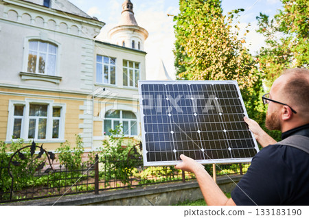 Man holding photovoltaic solar panel in front of historical building. Concept of integration of sustainable renewable energy sources into architecture. 133183190