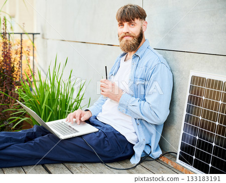 Portrait of barded man works on laptop on lap with glass of iced coffee. Solar panel nearby, emphasizing sustainable, modern eco-friendly workspace that combines technology and sustainability. Portrait of barded man works on laptop on lap with glass of iced coffee. Solar panel nearby, emphasizing sustainable, modern eco-friendly workspace that combines technology and sustainability. 133183191