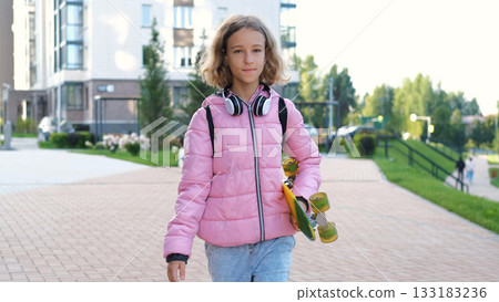 Teen girl walks to school in the morning carrying a colorful skateboard wearing a pink jacket and backpack with headphones around her neck Teen girl walks to school in the morning carrying a colorful skateboard wearing a pink jacket and backpack with headphones around her neck 133183236