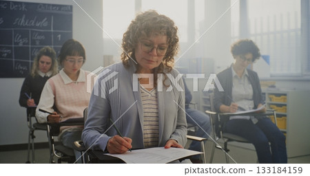Mature Woman With Curly, Greying Hair Sits at Desk, Deeply Focused on Written Test Among Students 133184159