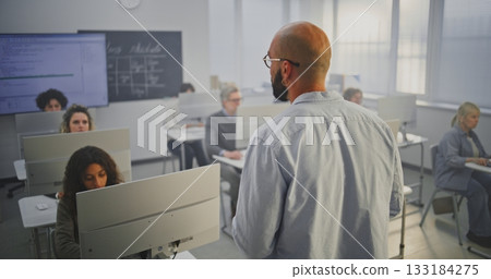 Professional Male Instructor Stands in Front of Adult Students in Computer Lab, Explaining Coding 133184275