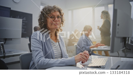 Cheerful Middle Aged Woman With Curly Graying Hair Studies Coding on Computer in Modern Classroom Cheerful Middle Aged Woman With Curly Graying Hair Studies Coding on Computer in Modern Classroom 133184318