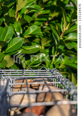 A vertical shot of a gabion against a green bush, an idea for decoration or fencing in landscape design. A vertical shot of a gabion against a green bush, an idea for decoration or fencing in landscape design. 133184476