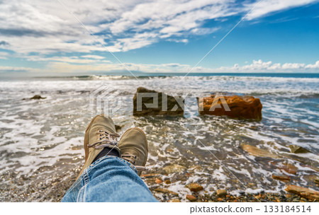 Feet in sneakers against the backdrop of the surf and a rocky seashore. A magnificent view and space for rest and relaxation after the work week, weekend by the sea concept 133184514