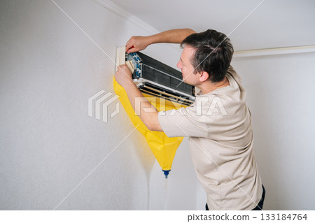 Skilled HVAC technician carefully disassembling air conditioning unit, using protective bag to catch dripping water and debris during repair process. Concept of air condition maintenance. Skilled HVAC technician carefully disassembling air conditioning unit, using protective bag to catch dripping water and debris during repair process. Concept of air condition maintenance. 133184764