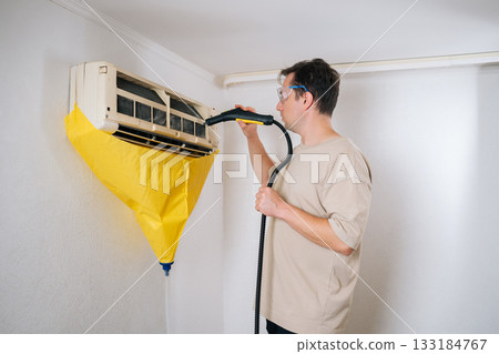 Side view of air conditioning technician cleaning air conditioner unit using steam cleaner. While yellow protective bag collects dirty water. Ensuring thorough and professional cleaning process. Side view of air conditioning technician cleaning air conditioner unit using steam cleaner. While yellow protective bag collects dirty water. Ensuring thorough and professional cleaning process. 133184767