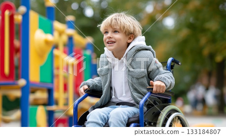 Blonde caucasian young boy in wheelchair smiling at colorful playground outdoors. 133184807