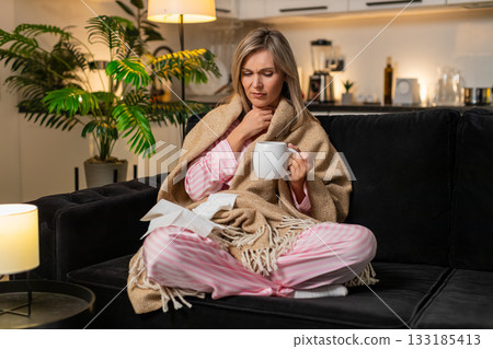 Adult woman sick with sore throat in blanket holds tea takes sips wipes nose with tissues at night 133185413