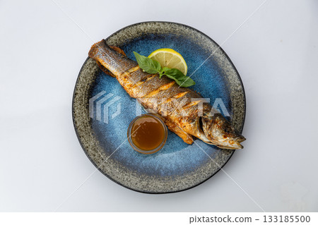 Whole grilled sea bass with lemon, basil and sauce on a blue ceramic plate, top view 133185500
