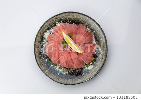 Tuna carpaccio with lemon and parmesan on greens, black plate, studio top view 133185503