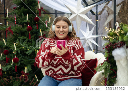 Cute young smiling woman listening music from smartphone during cozy Xmas holidays at home. Smiling Caucasian millennial lady in headphones sits near a Christmas tree Cute young smiling woman listening music from smartphone during cozy Xmas holidays at home. Smiling Caucasian millennial lady in headphones sits near a Christmas tree 133186932