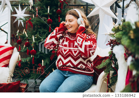 Cute young smiling woman listening music from smartphone during cozy Xmas holidays at home. Smiling Caucasian millennial lady in headphones sits near a Christmas tree Cute young smiling woman listening music from smartphone during cozy Xmas holidays at home. Smiling Caucasian millennial lady in headphones sits near a Christmas tree 133186942