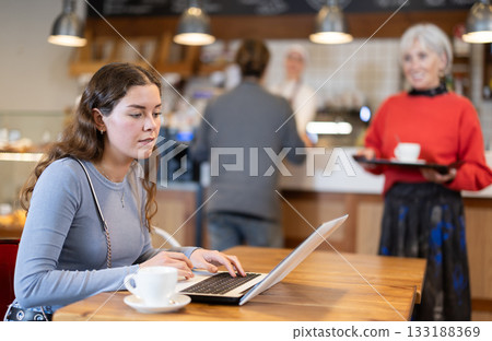 Female freelancer sitting at table in cafe with cup of coffee and laptop, browsing websites on internet 133188369