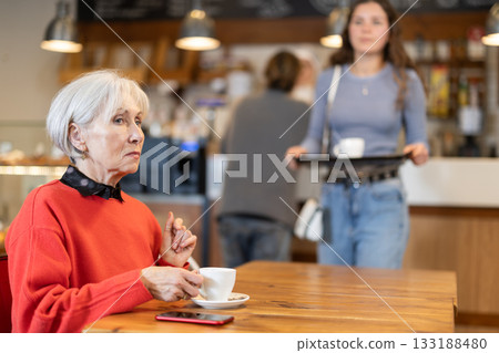 Mature woman with cup in hands drinking coffee in coffee shop indoors Mature woman with cup in hands drinking coffee in coffee shop indoors 133188480