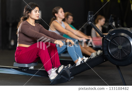Sportive Latina girl training with rowing machine in sports hall Sportive Latina girl training with rowing machine in sports hall 133188482