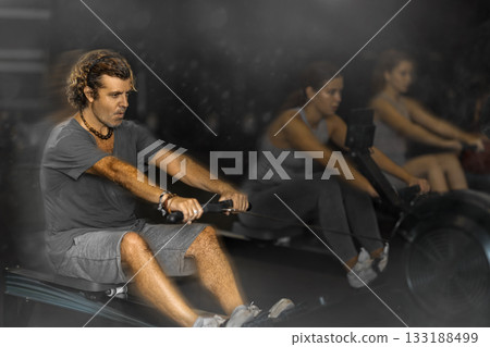 Diligent middle-aged man training with rowing machine in sports hall Diligent middle-aged man training with rowing machine in sports hall 133188499