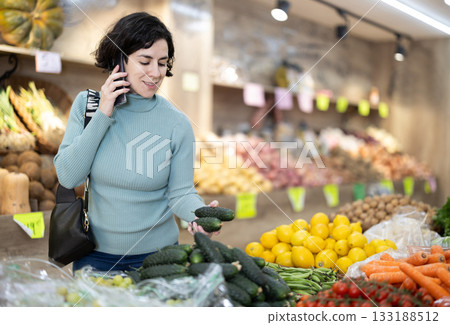 Adult woman choosing cucumbers while talking on phone Adult woman choosing cucumbers while talking on phone 133188512