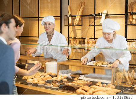 Two young old female bakery sellers helping with choice of delicious baked goods for customers behind counter in local bakeshop Two young old female bakery sellers helping with choice of delicious baked goods for customers behind counter in local bakeshop 133188633