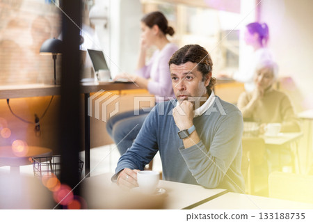 Serious adult male in casual wear sitting at table and enjoying coffee or tea in cozy cafeteria 133188735