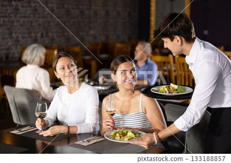 Young male waiter serving dish to two women in restaurant Young male waiter serving dish to two women in restaurant 133188857