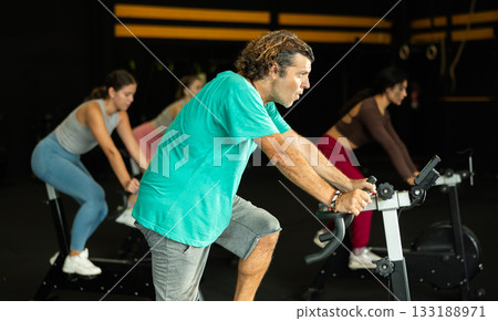 Diligent middle-aged man doing spin bike exercises in well-equipped gym Diligent middle-aged man doing spin bike exercises in well-equipped gym 133188971
