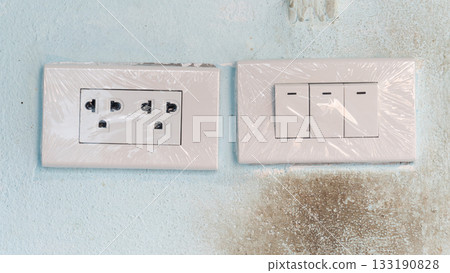 New electric plug three port with light switch three button. Cover confirm for new wrapped in plastic. On old cement wall blue color with stains. New electric plug three port with light switch three button. Cover confirm for new wrapped in plastic. On old cement wall blue color with stains. 133190828