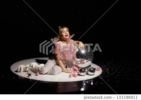 Girl in pink top and sequin pants sitting on stage with disco balls and dark background 133190911