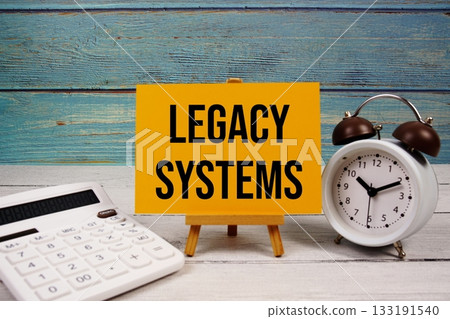 Legacy Systems text written on paper card with calculator and alarm clock on wooden background 133191540