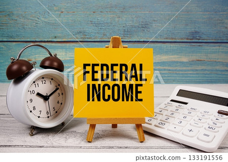 Federal Income text written on paper card with calculator and alarm clock on wooden background Federal Income text written on paper card with calculator and alarm clock on wooden background 133191556