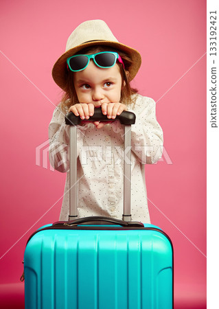 Bored little girl with blue suitcase, wears a straw hat and sunglasses, expresses puzzlement and confusion, portrait of child on isolated pink isolated. Bored little girl with blue suitcase, wears a straw hat and sunglasses, expresses puzzlement and confusion, portrait of child on isolated pink isolated. 133192421