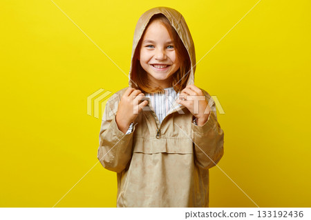 Cheerful portrait of happy little girl in demi-season beige jacket with hood from rain, studio shot on colored background. 133192436