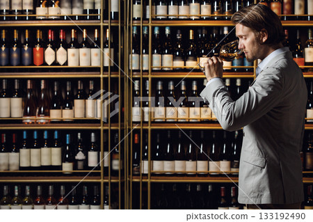 Indoor portrait of experienced male sommelier smells wine in glass standing in cellar over plenty bottle shelf background with a free copy space for your text or logo. Sommelier exam to study wine. Indoor portrait of experienced male sommelier smells wine in glass standing in cellar over plenty bottle shelf background with a free copy space for your text or logo. Sommelier exam to study wine. 133192490