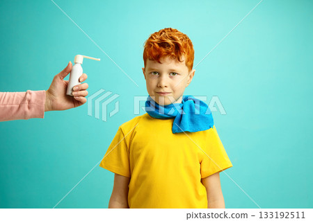 Seasonal Colds in Children Concept. Little Red Headed Boy Wrapped in a Scarf Preparing to Take Cough Throat Spray From Hands of Adult Standing on Blue Isolated Background 133192511