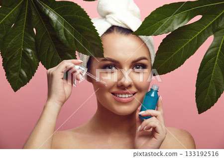 Smiling beautiful young woman in head towel holding facial serum stands among tropical leaves over pink isolated background. Advertisement of beauty product. 133192516