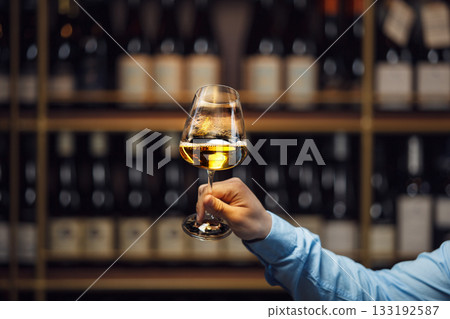 Expert Evaluating White Wine Glass Against Blurred Wine Cellar Background Expert Evaluating White Wine Glass Against Blurred Wine Cellar Background 133192587