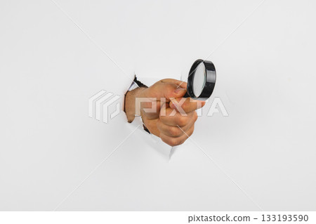 Hand holding magnifying glass emerging from a hole in the wall Hand holding magnifying glass emerging from a hole in the wall 133193590